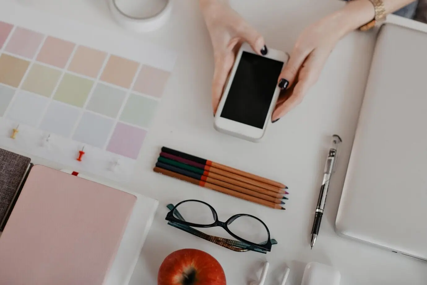 A snapshot of devices, a notepad, glasses, and apples on a white table.